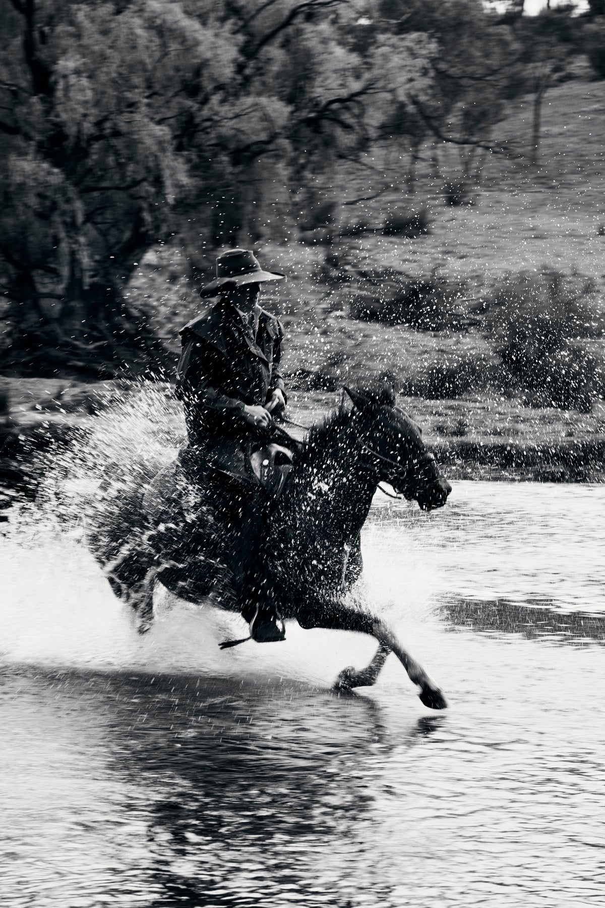 Black and white photograph of a cowboy riding a horse through splashing water, with trees and grass in the background, wearing the Driza-Bone Heavyweight Rider Coat.