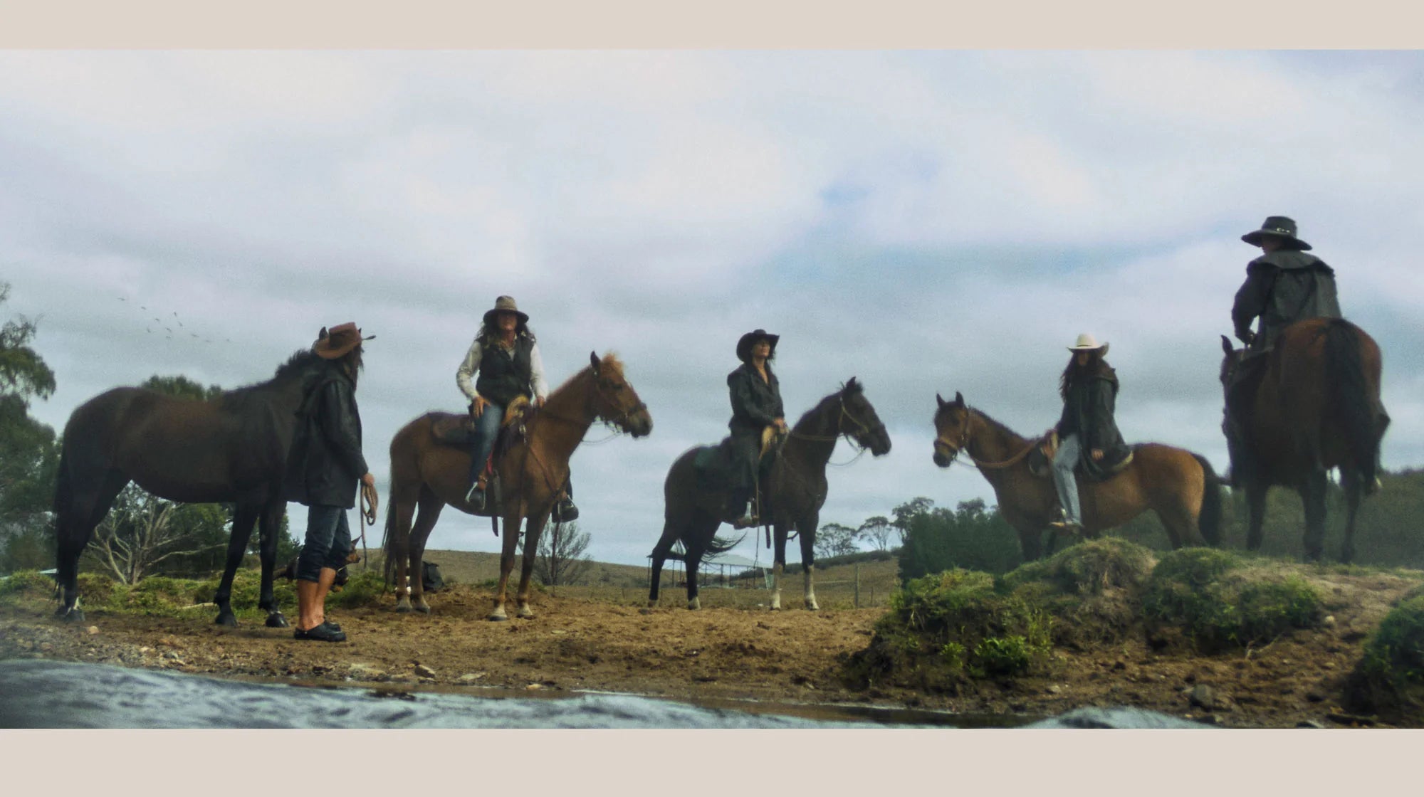 Five people on horseback in a line on a grassy field with a cloudy sky.