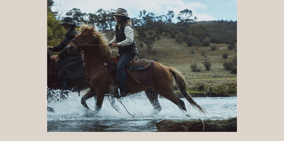 Two people riding horses through a river with a scenic landscape in the background, wearing Driza-Bone Heavyweight jackets and vests.