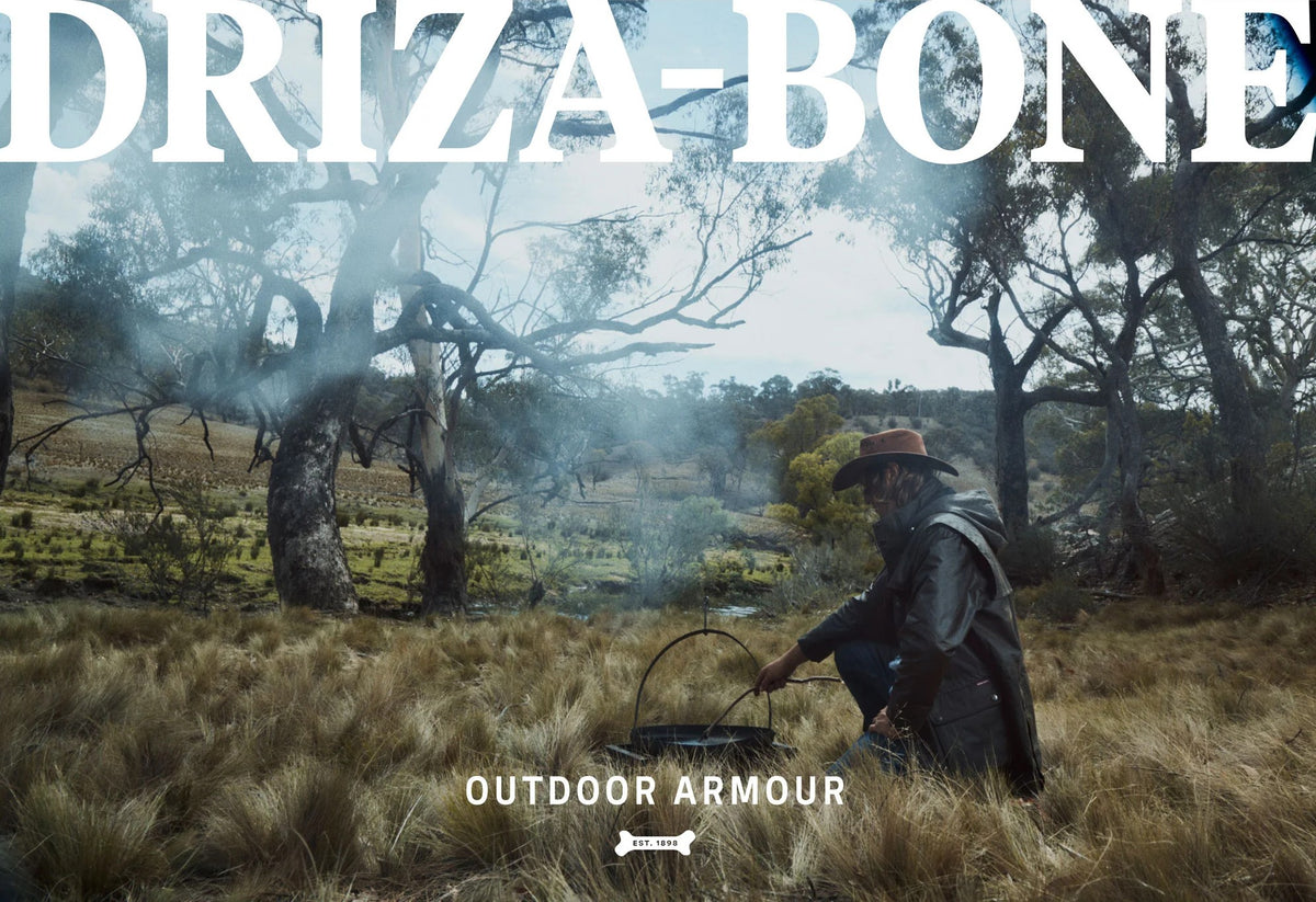 Person in outdoor setting with text overlay stating 'DRIZA-BONE' and 'Outdoor Armour' displayed.
Driza-Bone Dogbone logo visible at bottom of image. Man wearing the Driza-Bone Heritage Rider Coat.