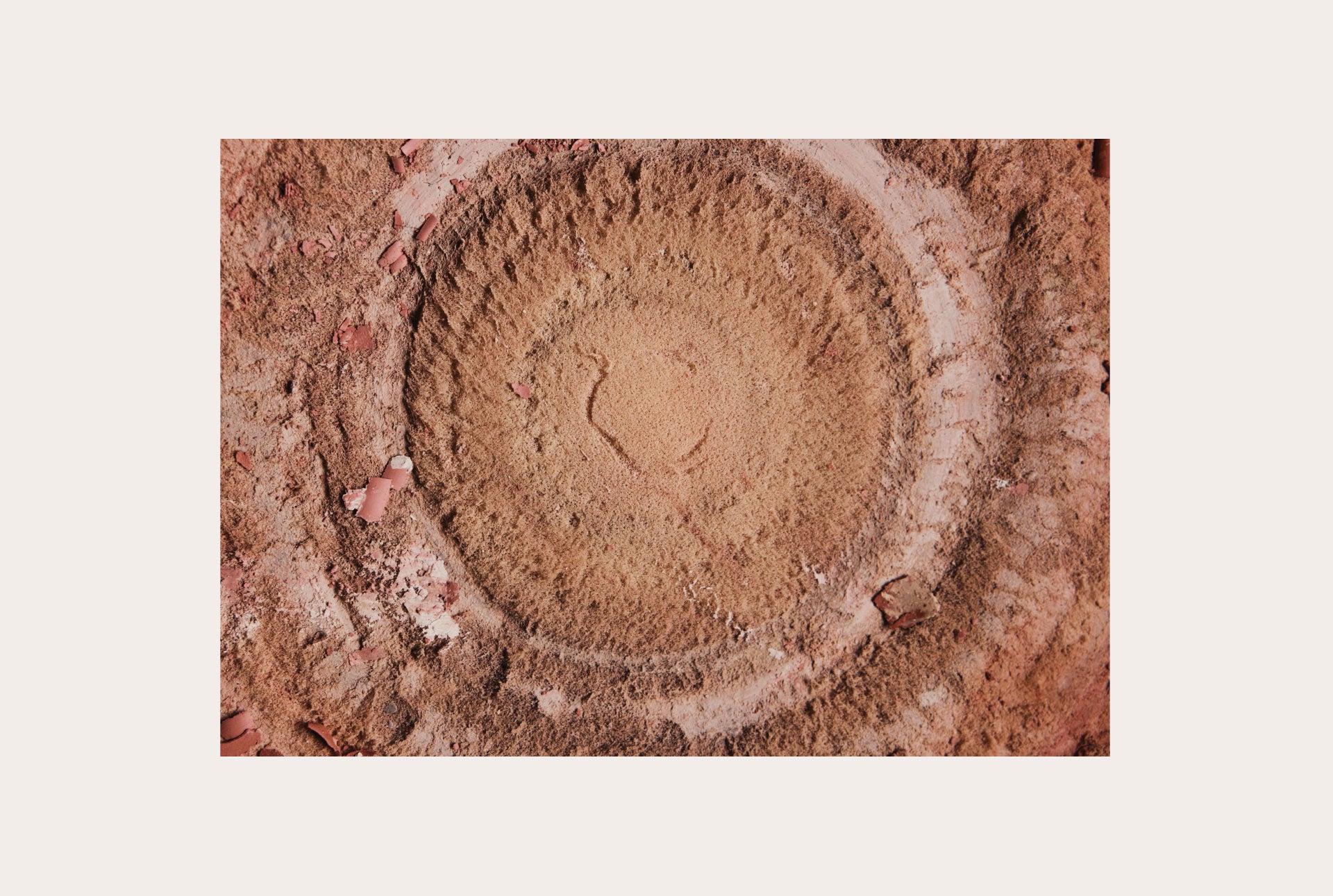A square art print featuring a top-down photograph of a circular, textured earthy brown surface, presented on a light beige background.