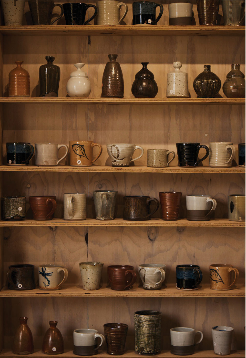 A collection of handmade ceramic mugs and small pottery vessels displayed on wooden shelves in a warm, rustic setting.