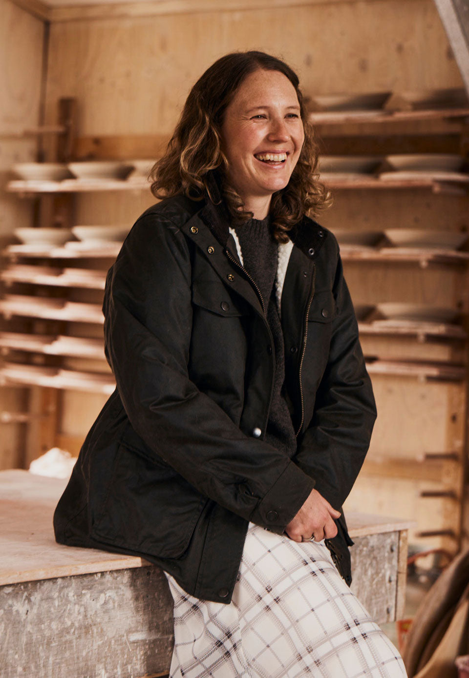 A woman with shoulder-length brown hair sits smiling on a wooden workbench inside a pottery or ceramic workshop. She is wearing the Driza-Bone Women's Lumberjack Jacket over a dark top and a white plaid skirt.