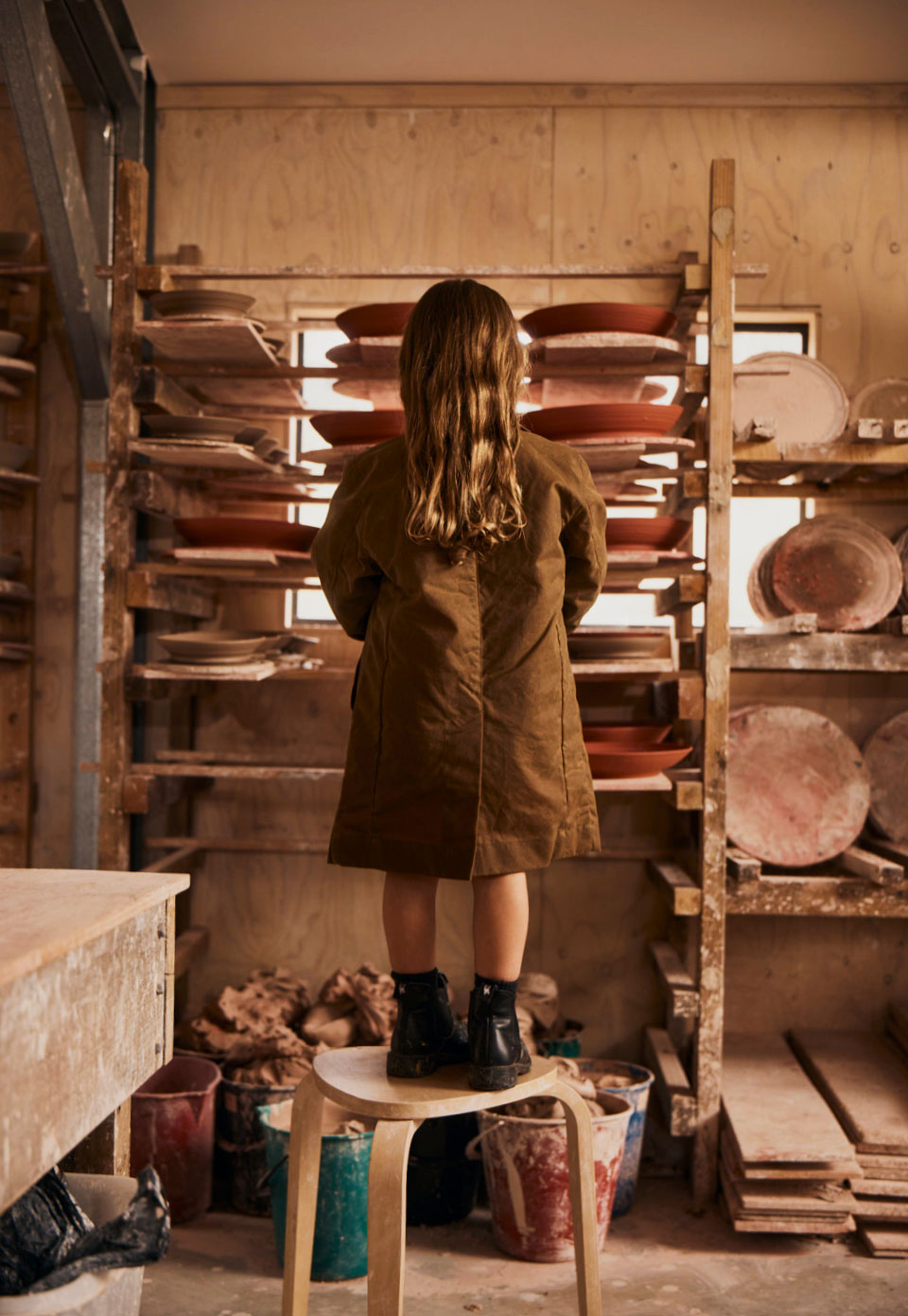 A child stands on a stool in a pottery studio, viewed from behind, wearing the Driza-Bone Kids Heavyweight Town and Country Coat in Tan colour and black boots, with shelves of clay plates and bowls in the background.