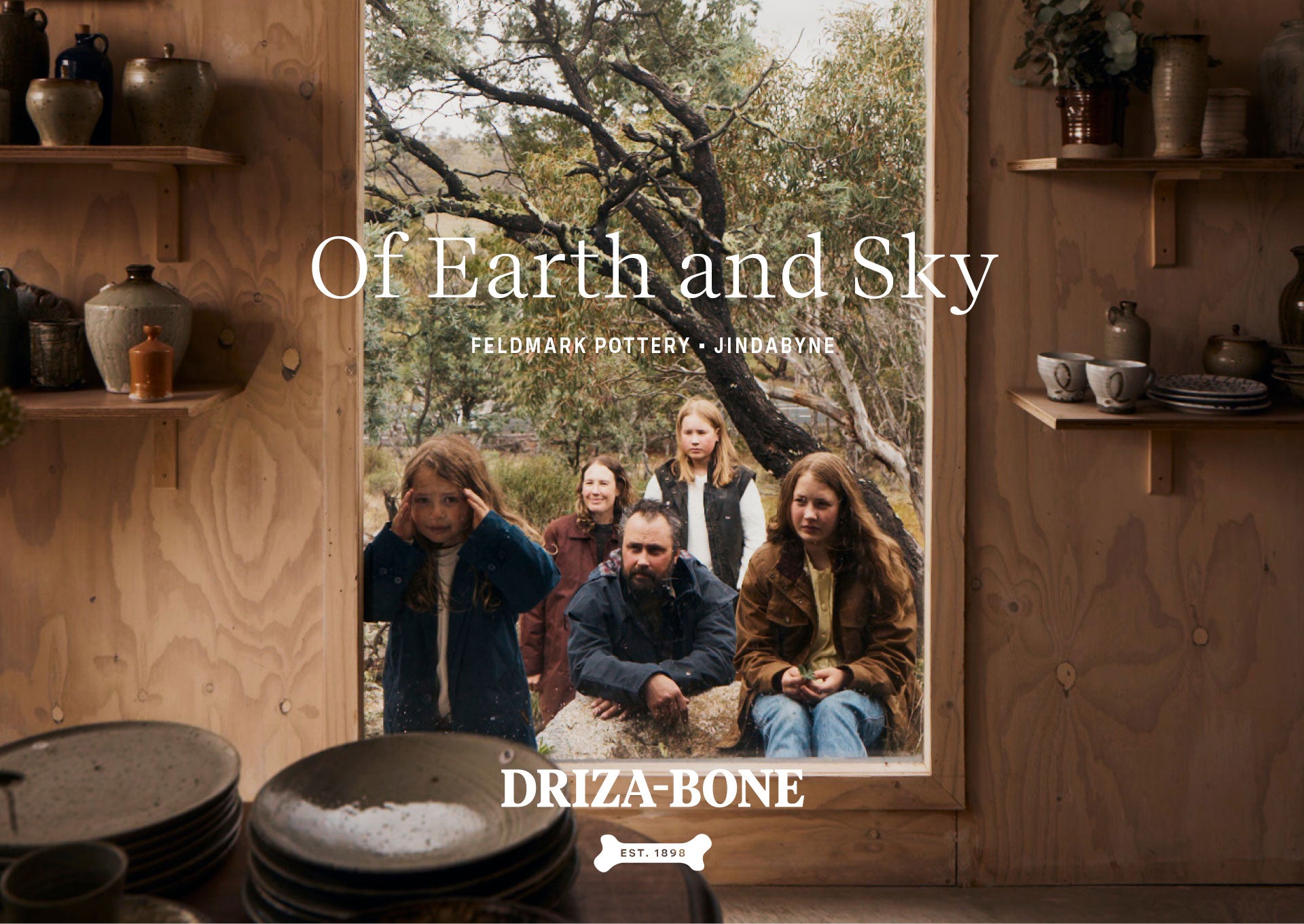 A rustic interior scene with wood-panelled walls, shelves holding ceramic pottery, and a large window showing four people outdoors among trees. Large overlaid text reads 'Of Earth and Sky' and 'FELDMARK POTTERY - JINJABYRE,' with 'DRIZA-BONE' at the bottom.