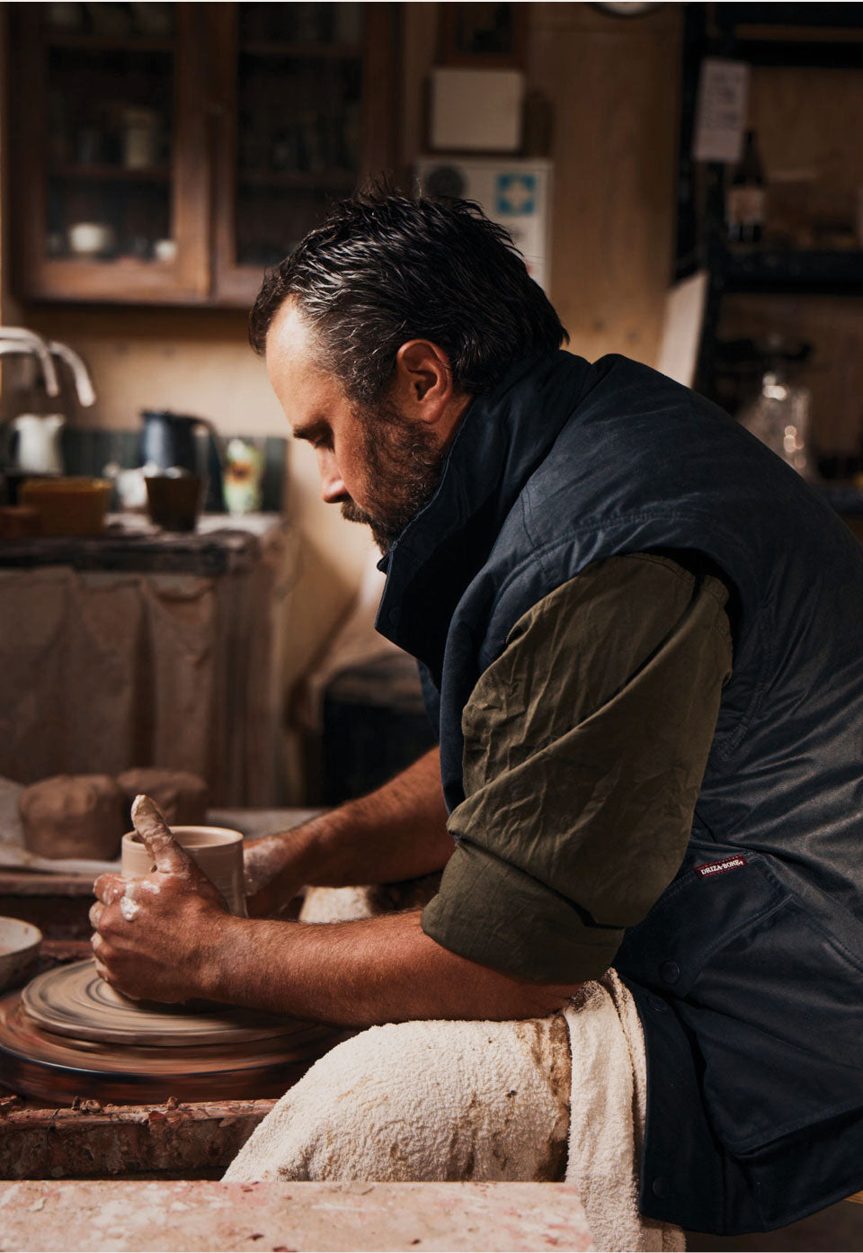 A man in a pottery workshop, shaping clay on a spinning wheel. He is wearing a dark navy quilted vest over an olive green shirt, with a white cloth around his waist.