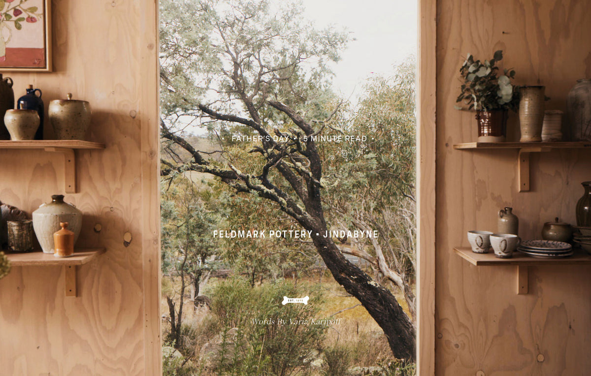 A rustic interior scene with light wood walls and shelves holding ceramic pottery, with a large window showing an outdoor tree view. White overlaid text reads 'FATHER'S DAY - 5 MINUTE READ' and 'FELMARK POTTERY - JINDBYNE', plus the Driza-Bone Dogbone logo near the bottom.